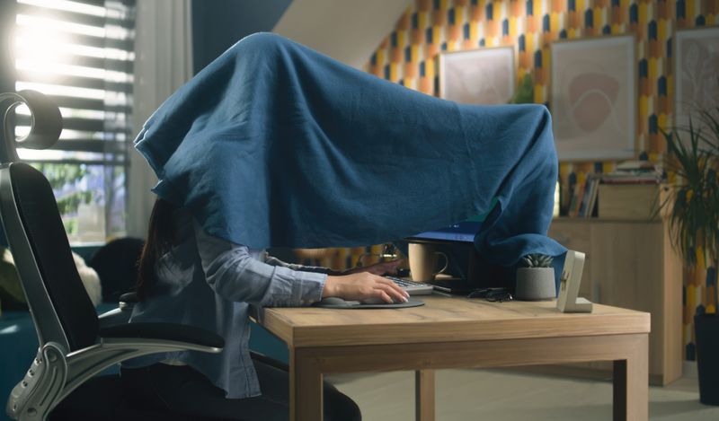 person at a desk hiding under a blanket